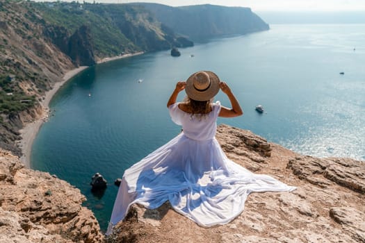 Woman sea. A happy girl is sitting with her back to the viewer in a white dress on top of a mountain against the background of the ocean and rocks in the sea
