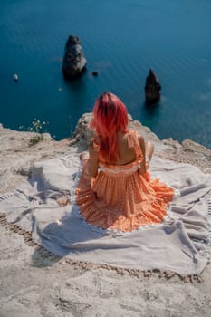 A girl with red hair sits with her back to the viewer on a picnic blanket in an orange dress. Summit on the mountain against the background of the sea and rocks in the sea