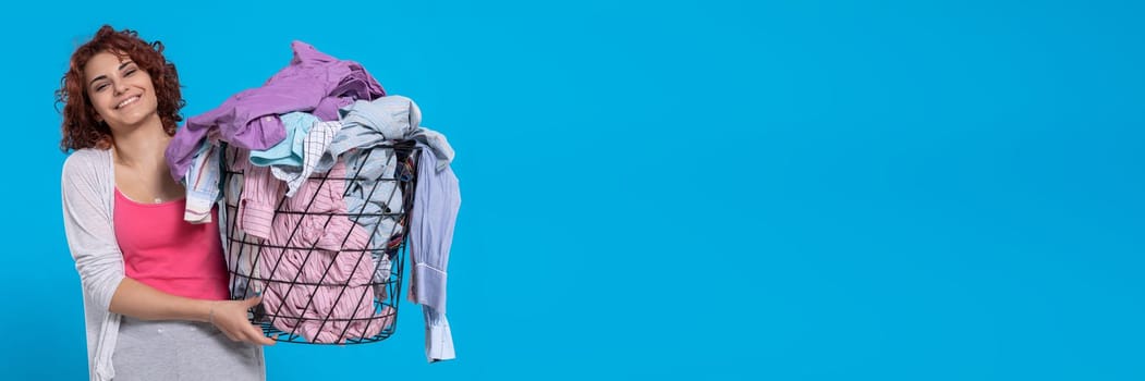 A girl with a smile on her face carries a basket full of washed and dried men's shirts. Joy and contentment. Dark curly hair. . Panoramic view of girl with empty blue background.