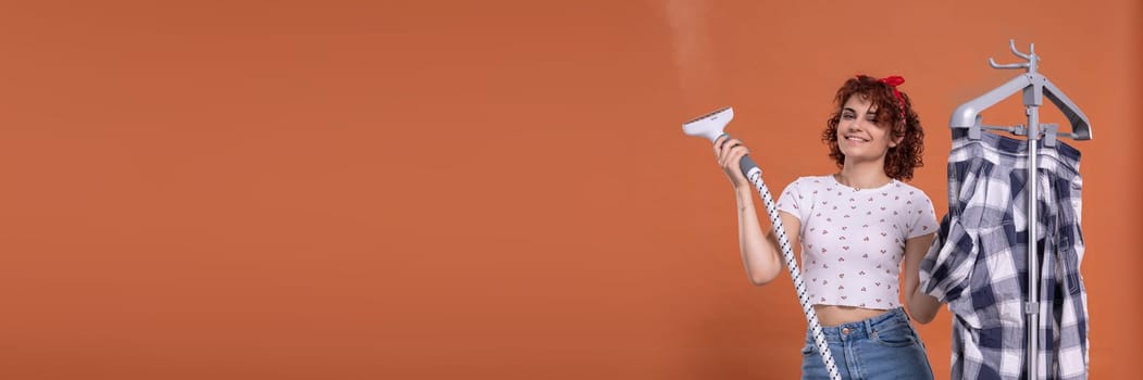 Hot steam used for ironing clothes. Steam device. An ironing steamer used by a young woman. Panoramic view of girl with empty orange background.
