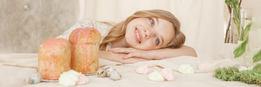 A girl with long hair in a light dress is sitting at the Easter table with cakes, spring flowers and quail eggs. Happy Easter celebration