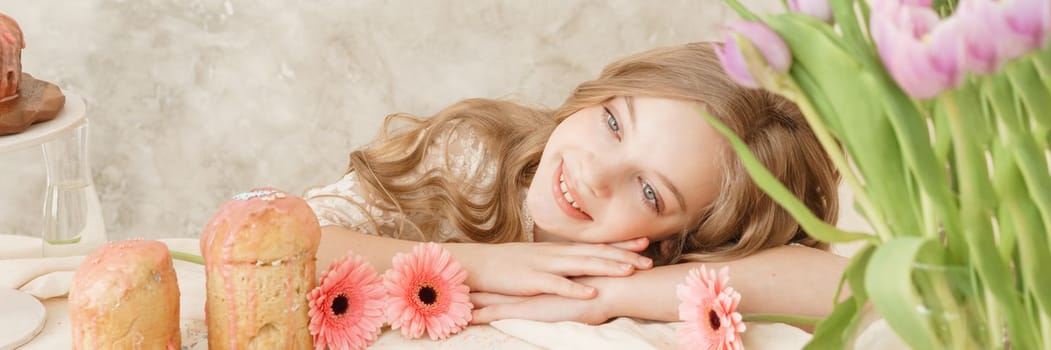 A girl with long hair in a light dress is sitting at the Easter table with cakes, spring flowers and quail eggs. Happy Easter celebration