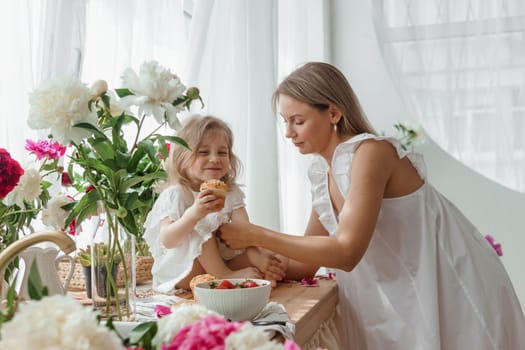 A little blonde girl with her mom on a kitchen countertop decorated with peonies. The concept of the relationship between mother and daughter. Spring atmosphere.