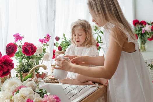 A little blonde girl with her mom on a kitchen countertop decorated with peonies. The concept of the relationship between mother and daughter. Spring atmosphere.