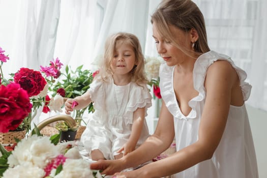 A little blonde girl with her mom on a kitchen countertop decorated with peonies. The concept of the relationship between mother and daughter. Spring atmosphere.