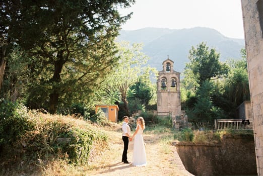 Bride and groom stand holding hands on the path to the ancient chapel. High quality photo