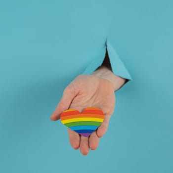 A hand with a rainbow-colored heart sticking out of a hole in a blue cardboard background