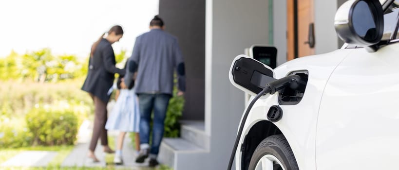 Focus closeup electric vehicle recharging battery from home electric charging station with blurred family in background. Renewable clean energy car for progressive eco awareness lifestyle concept.