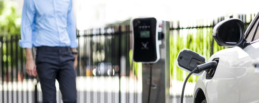 Focus closeup electric vehicle recharge battery at public charging station in the city area with blur businessman in background. EV car attached with electric charger for eco-friendly idea.