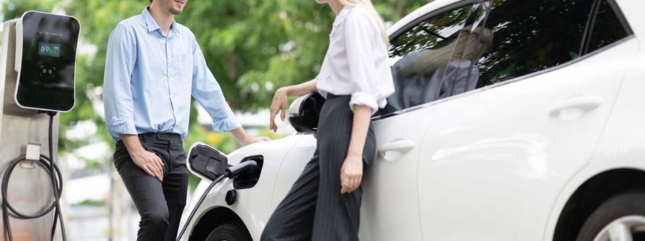 Progressive businessman and businesswoman with electric car parking and connected to public charging station before driving around city center. Eco friendly rechargeable car powered by clean energy.