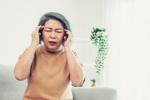 An agonizing senior woman with a headache, compress her temple with both hands at her contented living room. Senior health, nursing home, caretaker service.