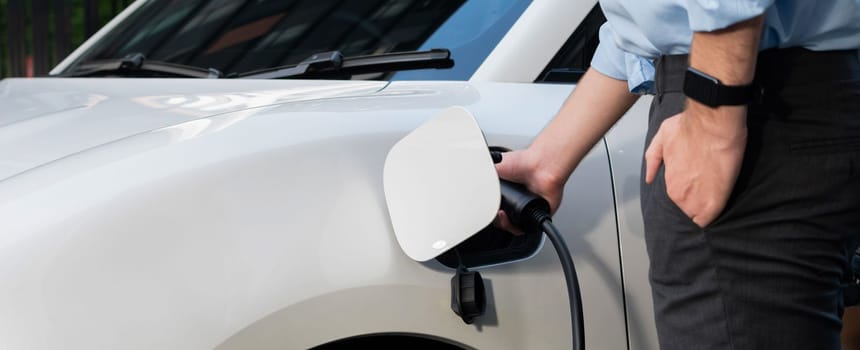 Closeup progressive man holding EV charger plug from public charging station for electric vehicle with background of residential building as concept eco-friendly sustainability energy car concept.