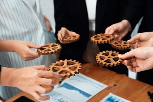 Closeup top view businesspeople hand holding gear and join together over meeting table with financial report papers. Cohesive group of office workers holding cog wheel as synergy harmony concept.