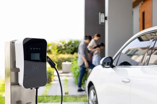 Focus closeup electric vehicle recharging battery from home electric charging station with blurred family in background. Renewable clean energy car for progressive eco awareness lifestyle concept.