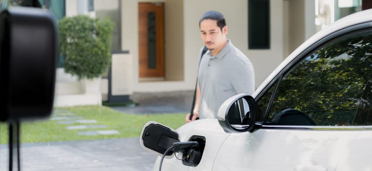 Progressive asian man install cable plug to his electric car with home charging station in the backyard. Concept use of electric vehicles in a progressive lifestyle contributes to clean environment.