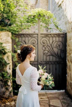 Bride with a bouquet of flowers stands in the courtyard of an old stone house. Back view. High quality photo