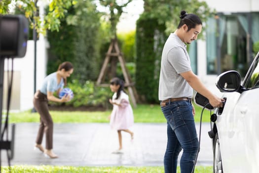 Focus image of progressive man charging electric car from home charging station with blur mother and daughter playing together in the background.