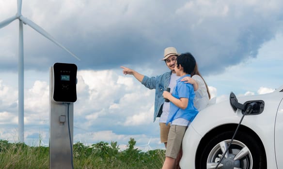 Concept of progressive happy family enjoying their time at wind farm with electric vehicle. Electric vehicle driven by clean renewable energy from wind turbine generator for charging station.