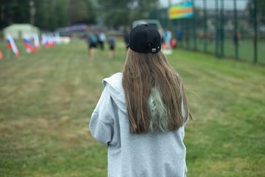 Girl in park. Teenager walks on street. Black cap and long hair. Grey jacket. Girl in summer.