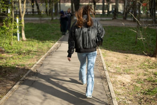 Girl walks down road. Woman in black jacket and blue jeans. Woman in park.