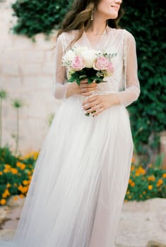 Bouquet of roses in the hands of a bride in a white dress standing in the yard. High quality photo