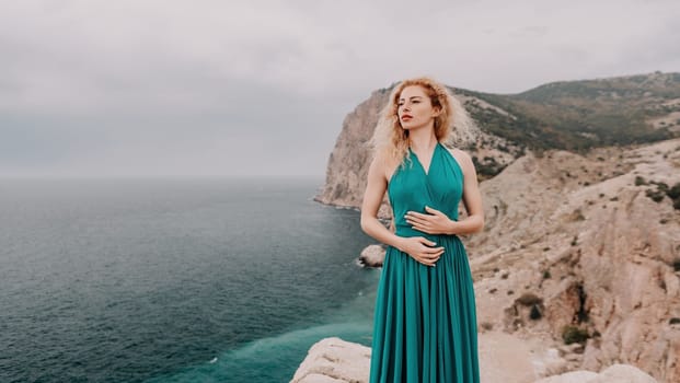 Side view a Young beautiful sensual woman in a mint long dress posing on a volcanic rock high above the sea during sunset. Girl on the nature on overcast sky background. Fashion photo