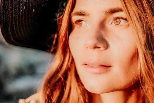 Portrait of happy young woman wearing summer black hat with large brim at beach on sunset. Closeup face of attractive girl with black straw hat. Happy young woman smiling and looking at camera at sea