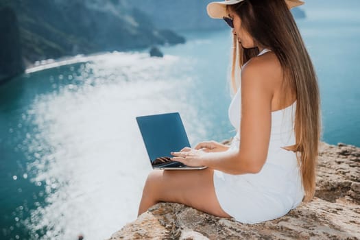 Successful business woman in yellow hat working on laptop by the sea. Pretty lady typing on computer at summer day outdoors. Freelance, travel and holidays concept.