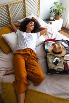Happy multiracial woman lying down on bed next to open suitcase full of clothes, camera, passport and hat, ready to go on summer vacation trip. Vertical image. Holiday and vacations concept.