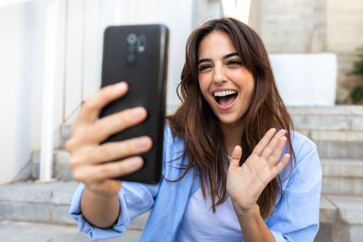 Happy young brunette woman waving hand during video call. Female influencer vlogger live streaming using phone sitting on stairs outdoors in street. Technology and lifestyle concept.