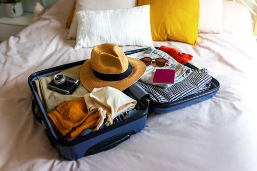 Open suitcase on the bed full of clothes and summer items: hat, sunglasses, swimsuit. Ready for vacations: passport and camera.