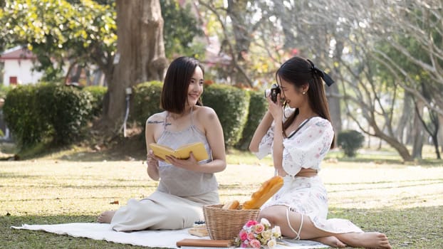 Two attractive young Asian women enjoying picnic in the park together, taking a photo from camera, reading a book.