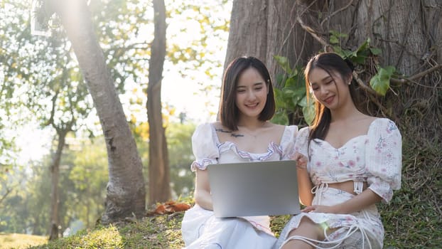 Two Asian women are watching a video on a laptop together while enjoying a picnic in the park.