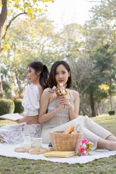 Charming beautiful young Asian woman in a lovely dress, holding a flower bouquet, enjoying a picnic with her friend in the park.