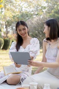Two young Asian women are using tablet, taking selfies with tablet or watching videos on a digital tablet together while enjoying a picnic in the park.