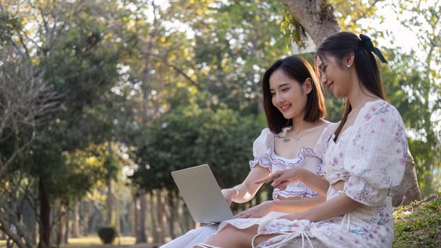Two Asian women are watching a video on a laptop together while enjoying a picnic in the park.