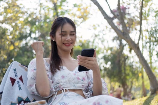 beautiful Asian woman using her smartphone while picnicking in the green park.