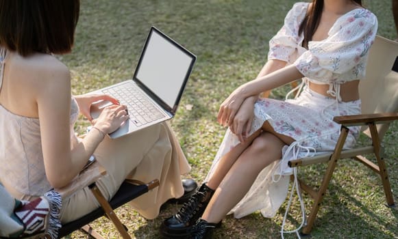 Two beautiful Asian women in lovely dresses enjoying picnic in the park, sipping , work on laptop sitting on picnic chairs.