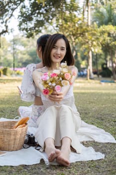 Charming beautiful young Asian woman in a lovely dress, holding a flower bouquet, enjoying a picnic with her friend in the park.