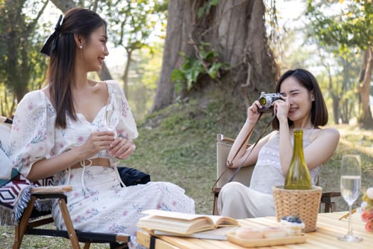 Charming Asian woman enjoying picnic in the park with her friend, taking photo with camera, having special moment on the weekend together.