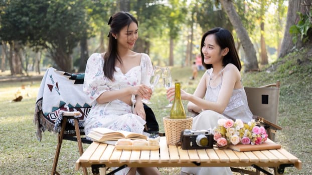 Two beautiful Asian women in lovely dresses enjoying picnic in the park, sipping white wine, sitting on picnic chairs.