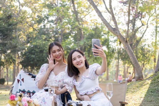 Two beautiful young Asian women picnic in the green park together, taking selfies with their phone they are having picnic,they talk happily.