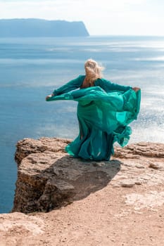 A beautiful young woman in a mint light dress with long legs stands on the edge of a cliff above the sea waving long dress, against the background of the blue sky and the sea