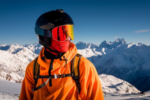 Portrait of a young man in a ski mask, stands in a ski resort against the backdrop of mountains and blue sky.