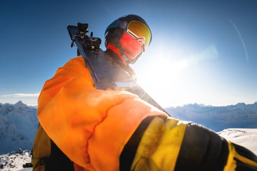 Portrait of a skier against the backdrop of the snow-capped mountains of the Caucasus. a man in a ski mask and a ski helmet on his shoulder holds skis and looks at the mountains. Ski resort concept.