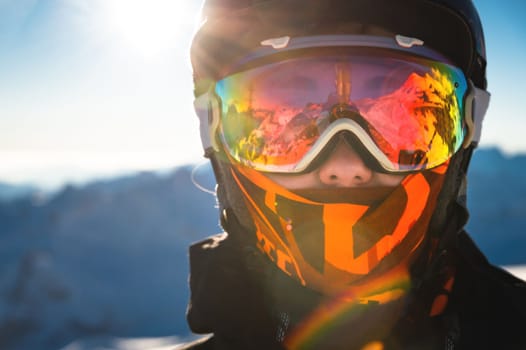 Portrait of a healthy woman in a sports mask looking directly at the camera against the backdrop of the sun and sunbeams, spending winter holidays in the snowy mountains.