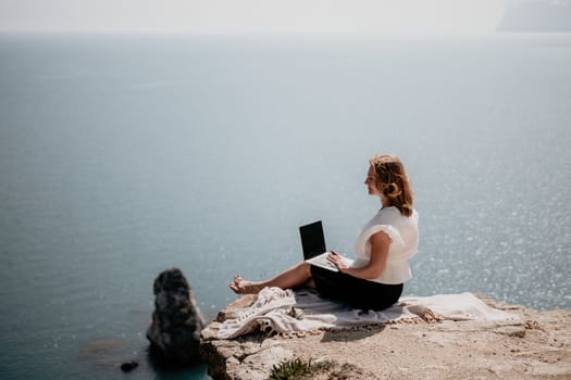 Successful business woman in yellow hat working on laptop by the sea. Pretty lady typing on computer at summer day outdoors. Freelance, travel and holidays concept.