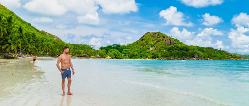 young man in swim short at a white tropical beach with turquoise colored ocean Anse Volbert beach Praslin Seychelles.
