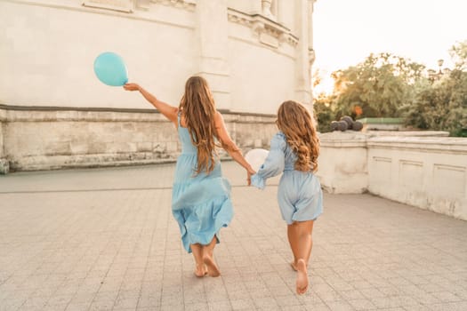 Daughter mother run holding hands. In blue dresses with flowing long hair, they hold balloons in their hands against the backdrop of a sunset and a white building