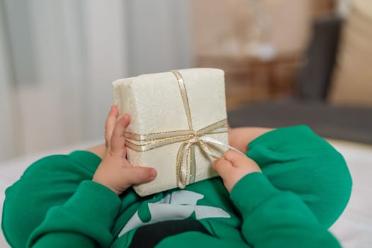 Kid gift. A little boy in a green suit holds a gift box with his feet and hands in bed